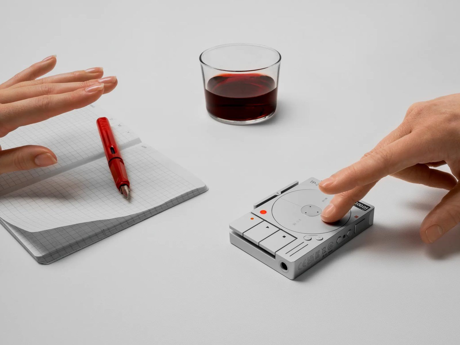 The TP-7 on a desk next to a notebook, a red fountain pen and a glass of red wine, a finger pressing a key