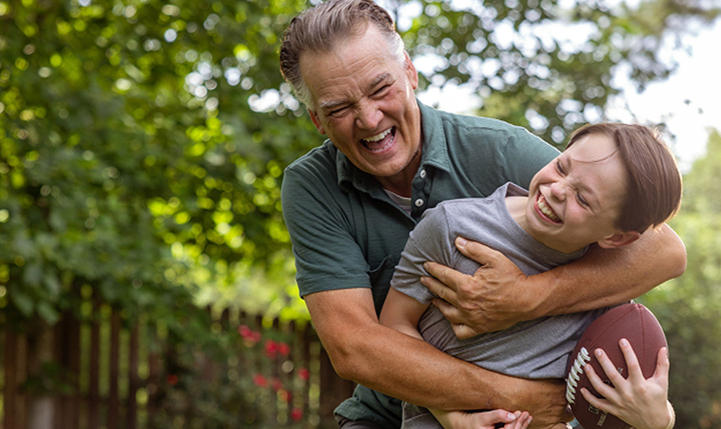 An older man and a young boy laughing and playfully wrestling with a football outdoors in a green yard.