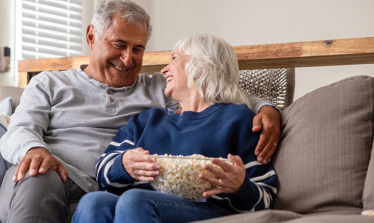 An older couple smiling and laughing together on a couch, with the woman holding a bowl of popcorn.