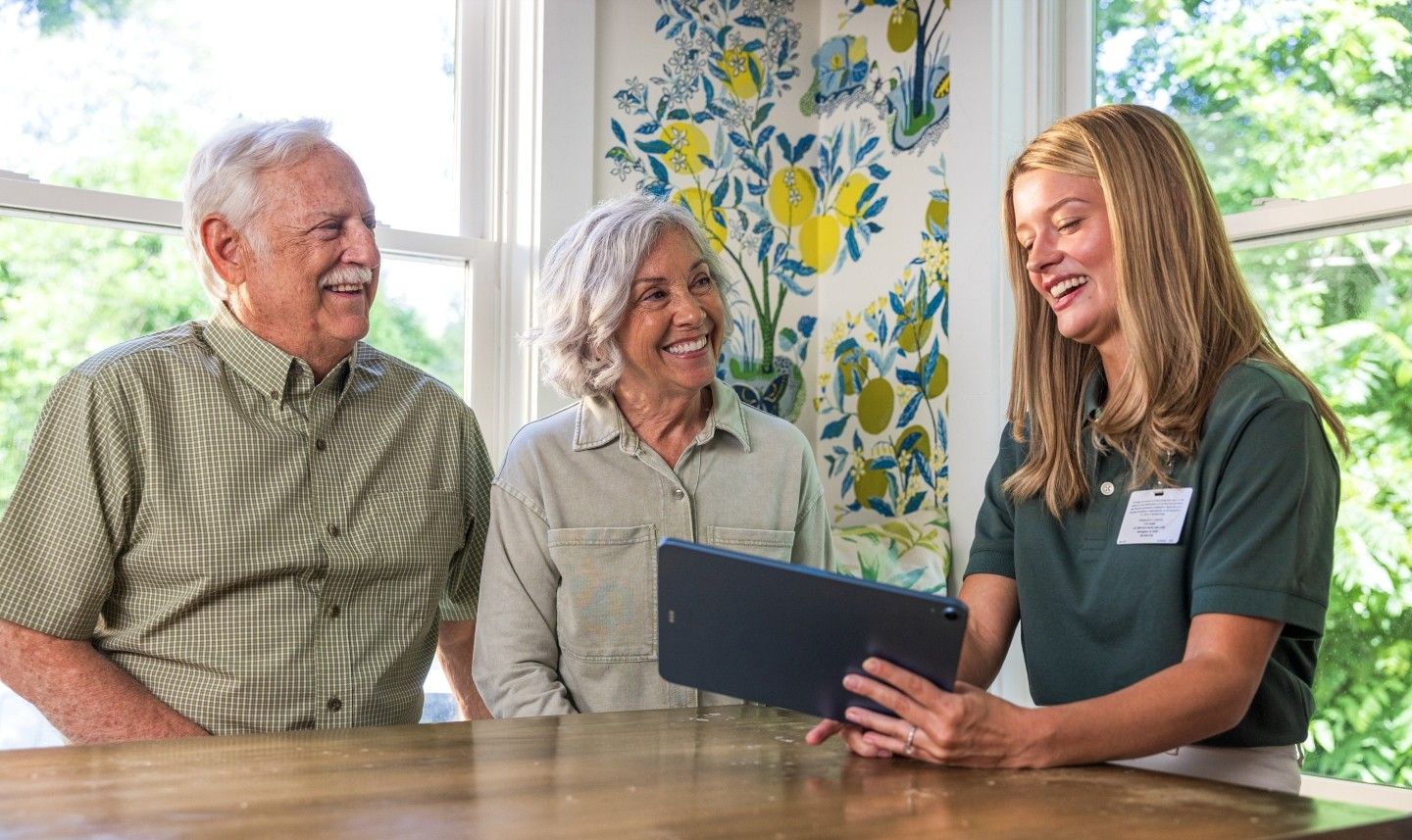A Viva Health representative showing a tablet to a smiling older couple sitting together at a wooden table.