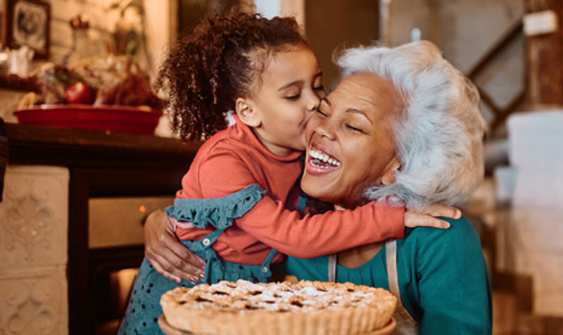 A young girl hugging and kissing a laughing older woman on the cheek in a warm kitchen, with a freshly baked pie on the counter in front of them.