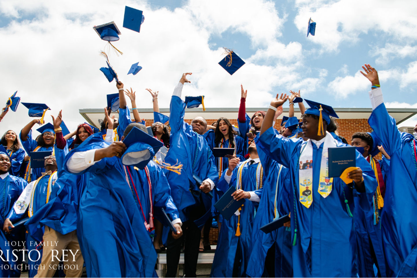 A group of graduating students from Holy Family Cristo Rey Catholic High School in blue gowns joyfully throwing their caps into the air.