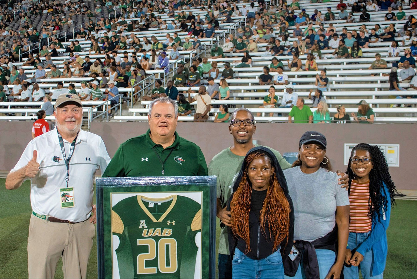 A group of six smiling people posing on a football field in front of a stadium crowd, with one man holding a framed green UAB football jersey.