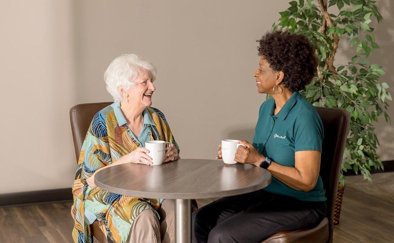 A woman in a green Viva Health polo shirt holding a coffee mug and conversing with a smiling older woman seated at a small round table.