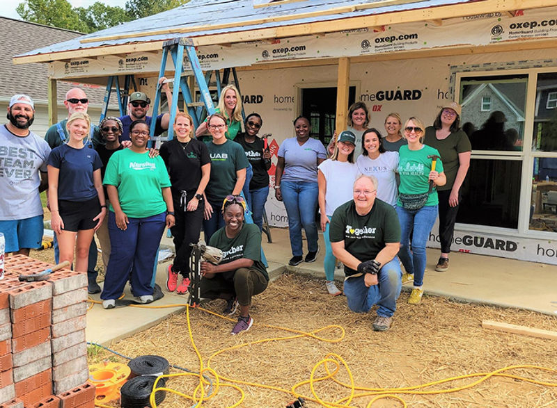 A large group of volunteers smiling in front of a partially constructed house.