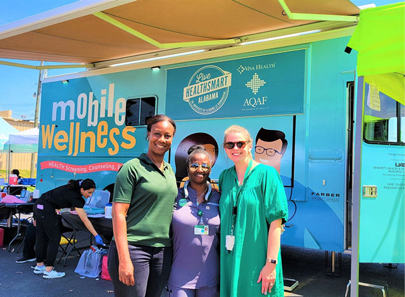 Three women posing outside in front of a bright blue Mobile Wellness clinic bus.