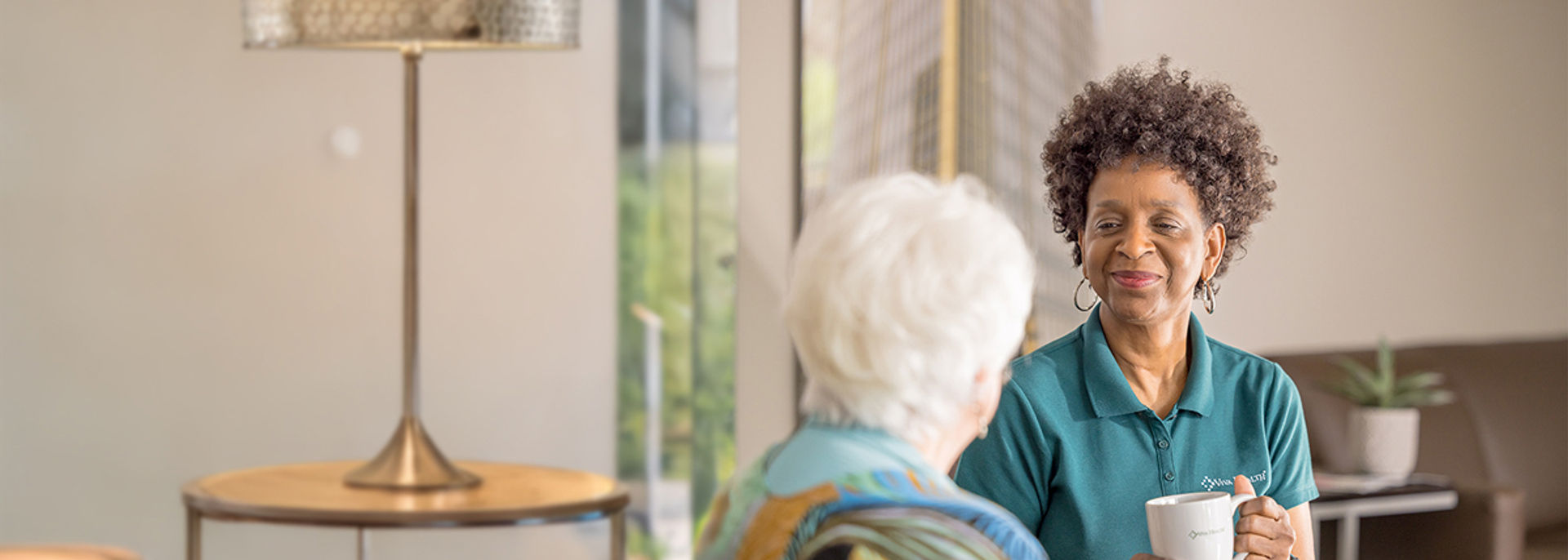 A smiling woman in a green Viva Health polo shirt holding a white coffee mug while speaking with an older woman in a brightly lit room.