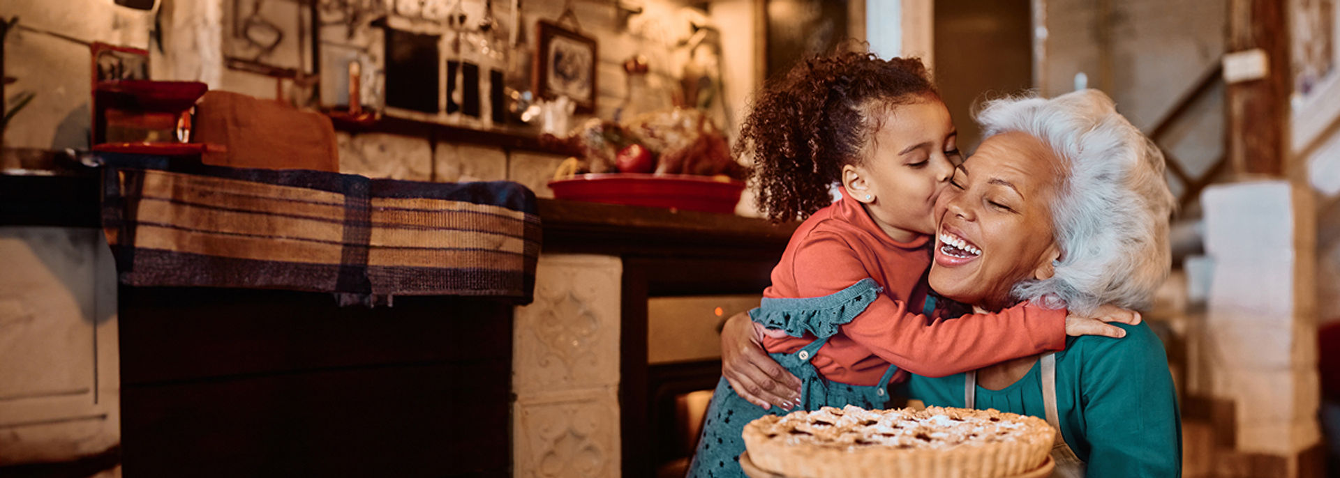A young girl hugging and kissing a laughing older woman on the cheek in a warm kitchen, with a freshly baked pie on the counter in front of them.