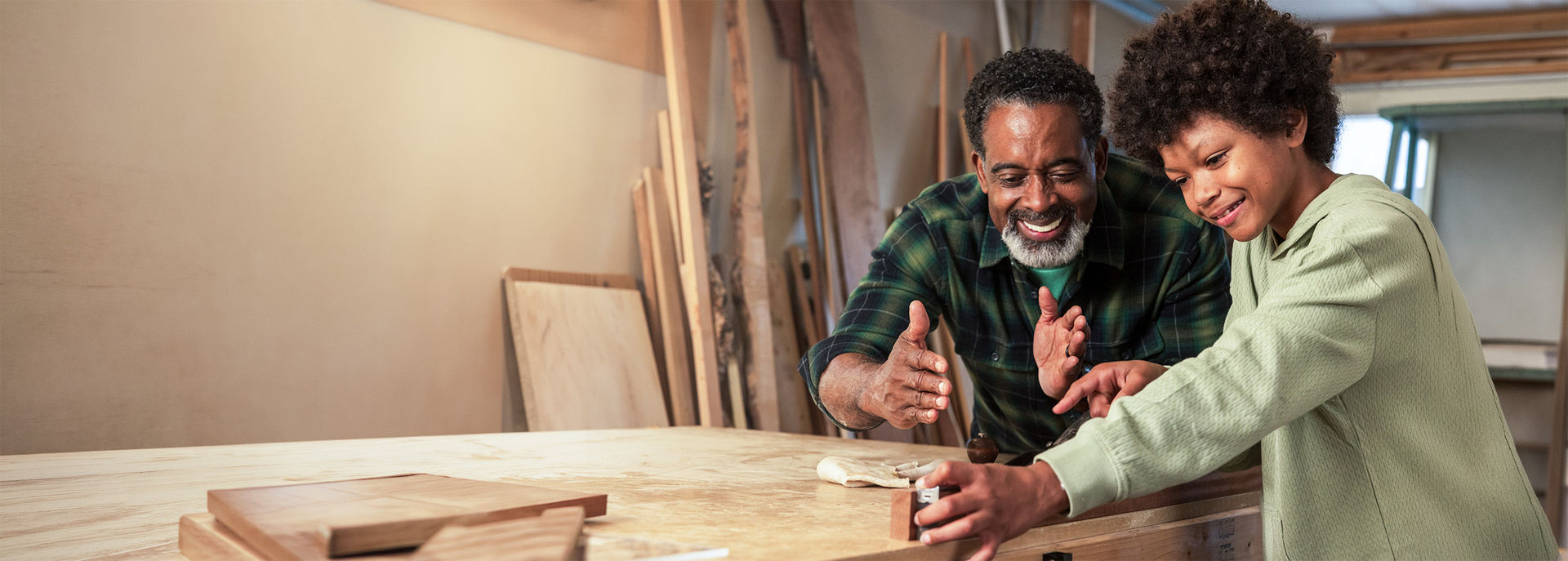 An older man smiling and guiding a young boy working on a woodworking project at a workbench in a workshop.