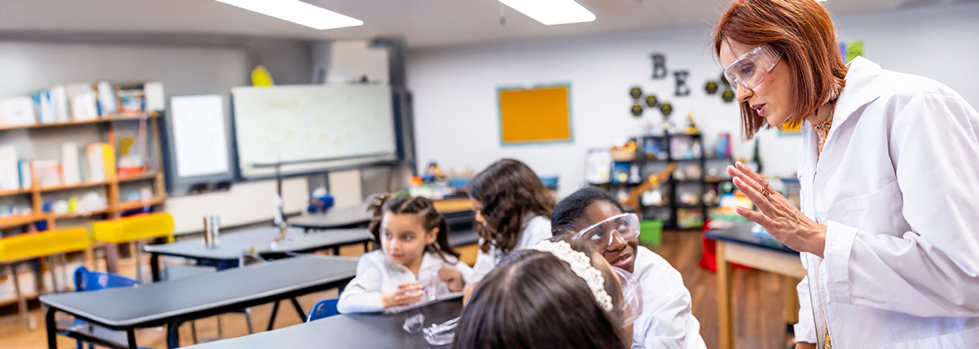 A female science teacher wearing a lab coat and safety goggles instructing young students in a classroom.