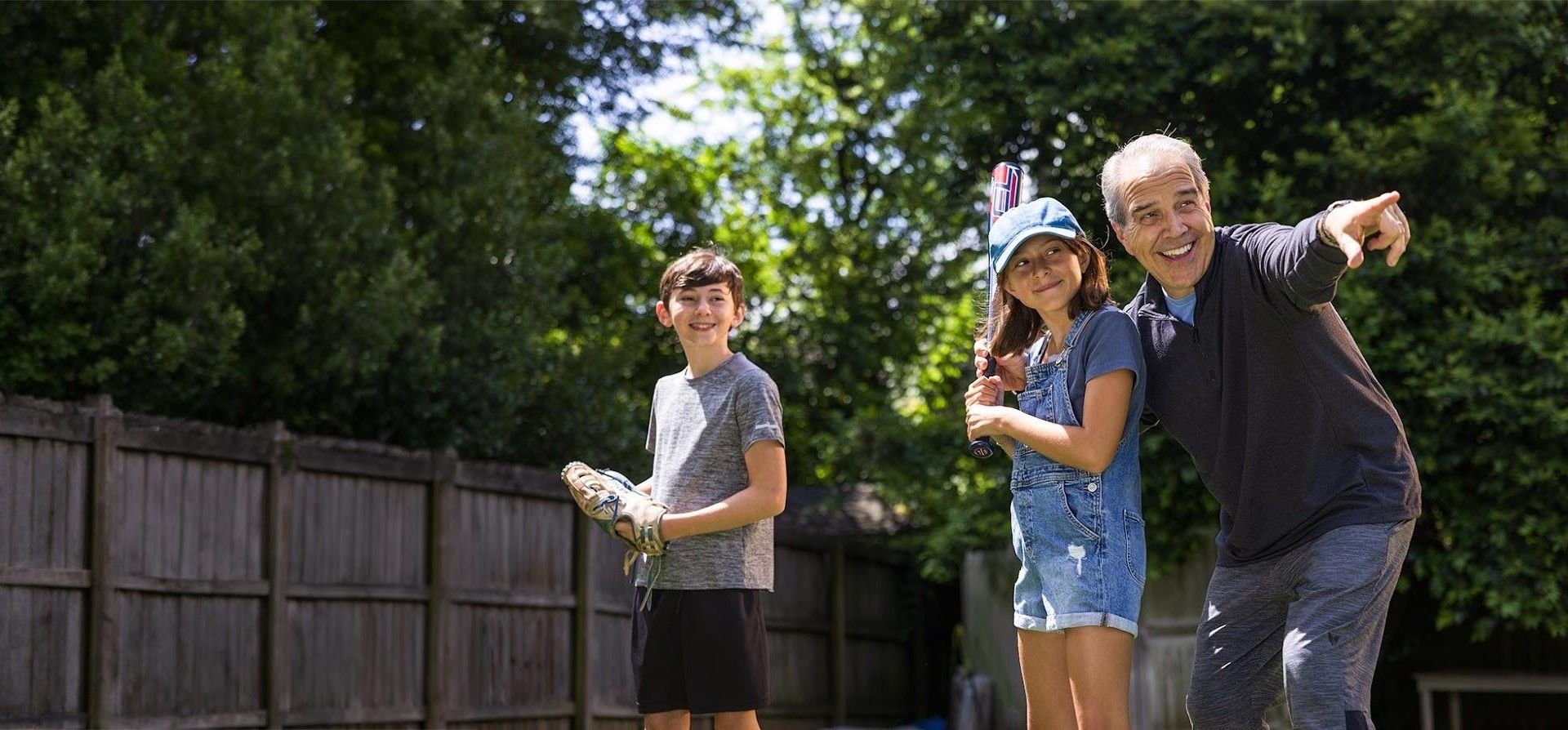 An older man pointing outward while playing baseball in a grassy yard with a young girl holding a bat and a boy holding a glove.