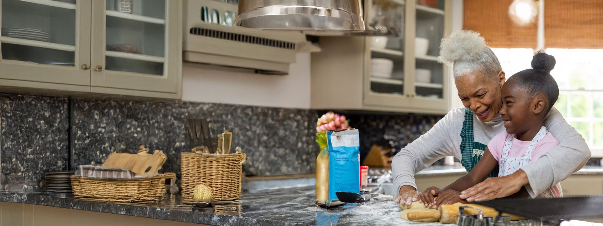 A smiling older woman and a young girl baking together in a kitchen, rolling out dough on a flour-covered countertop.