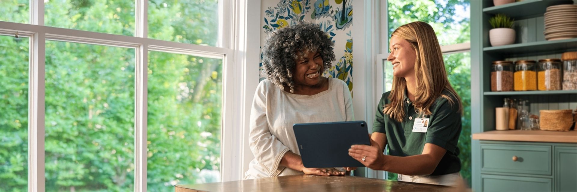 A smiling older woman and a female representative looking at a tablet together at a kitchen table.
