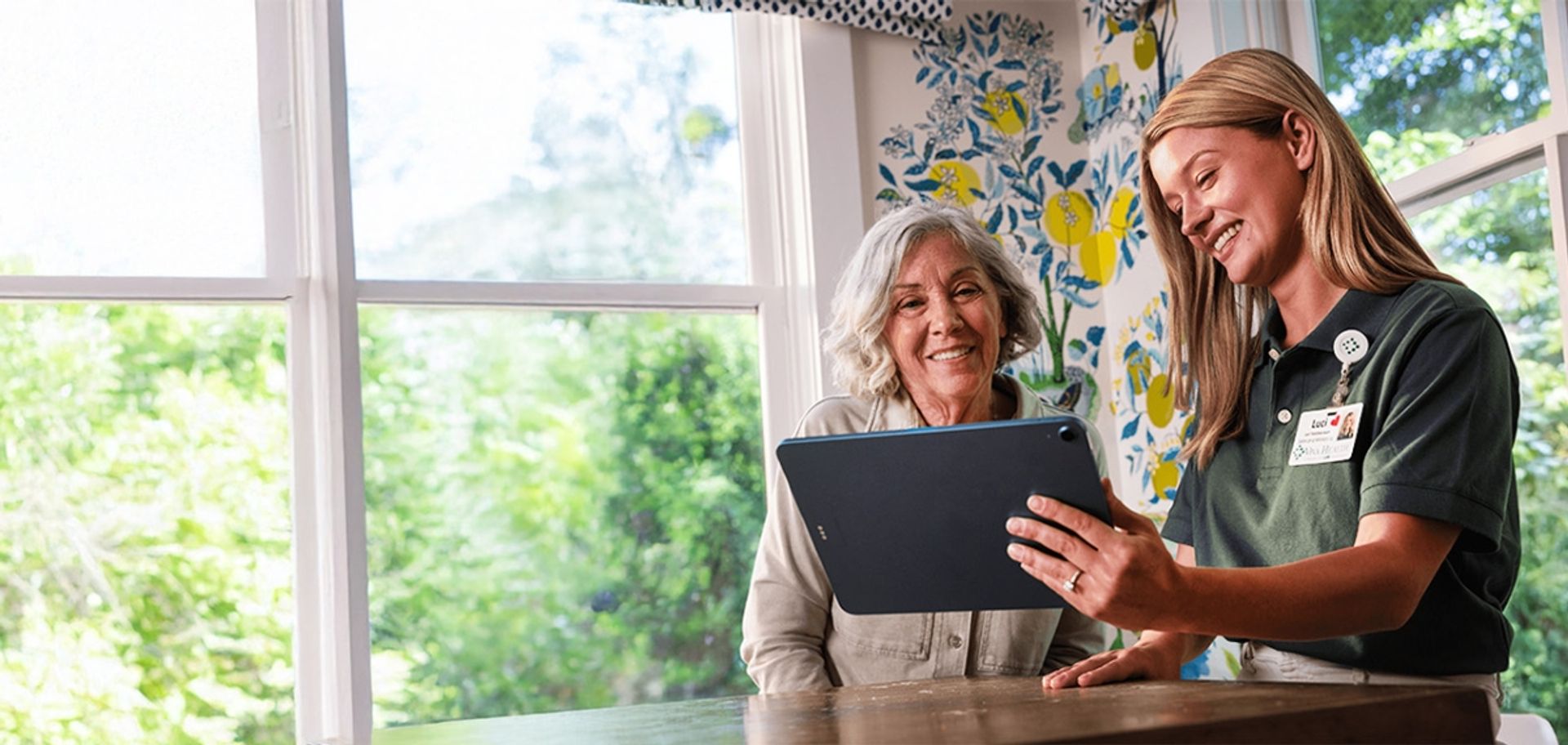 A smiling older woman and a female representative looking at a tablet together at a kitchen table.