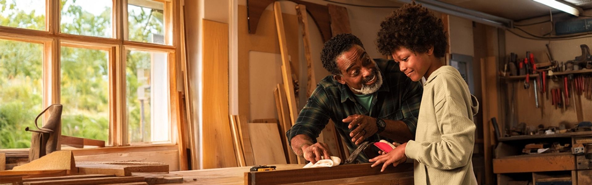 An older man and a young boy smiling as they work together on a woodworking project in a sunlit workshop.