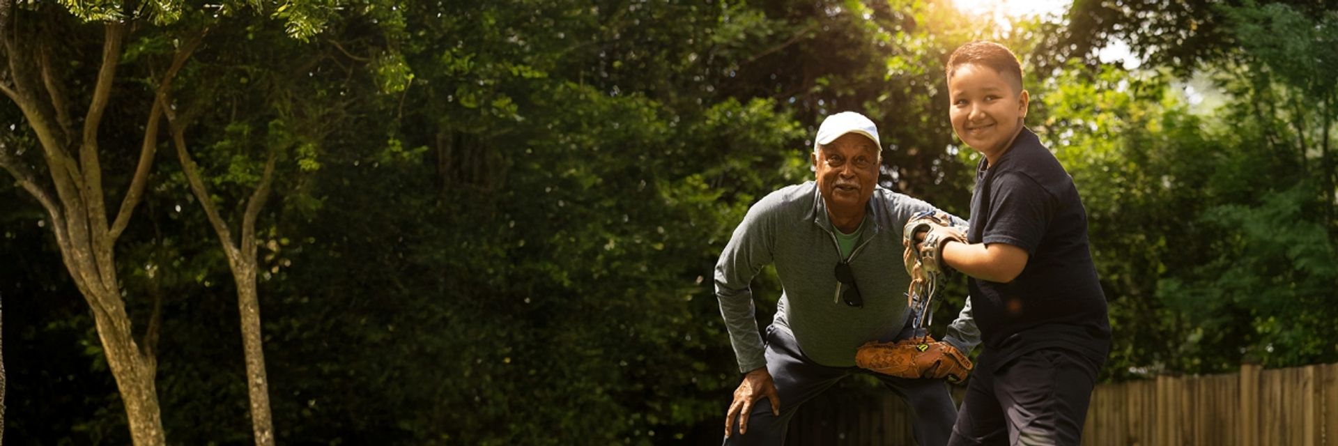A smiling older man and young boy wearing baseball gloves, playing together outdoors on a sunny day.