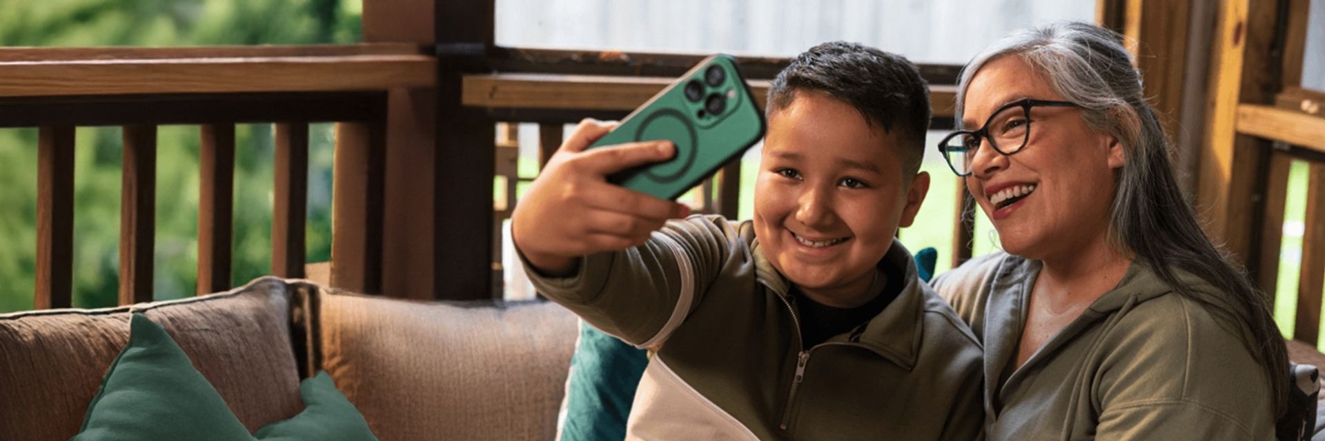 A smiling young boy holding a green smartphone to take a selfie with a happy older woman wearing glasses, sitting together on an outdoor porch sofa.
