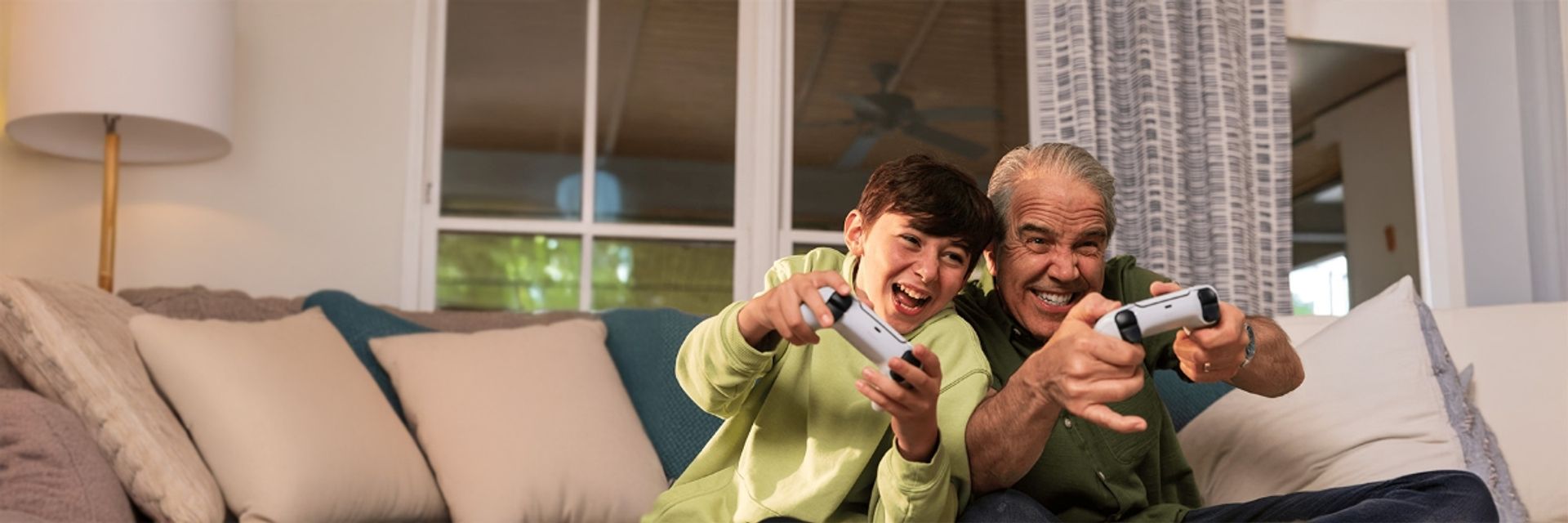 An excited young boy and an older man playing a video game together on a living room couch, both smiling widely and holding white controllers.