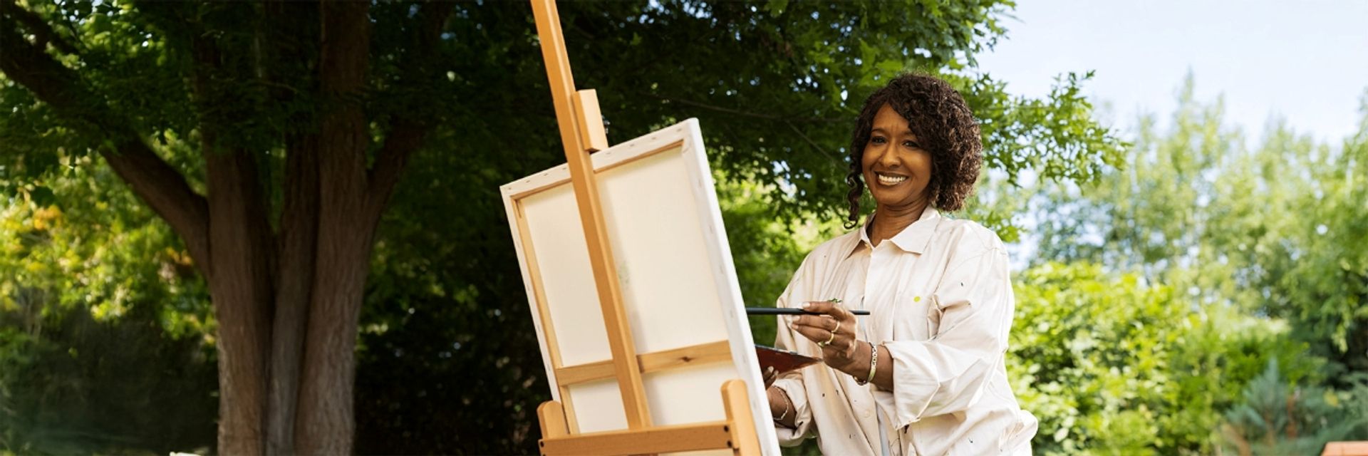 A smiling woman standing outdoors holding a paintbrush while painting on a canvas on a wooden easel.