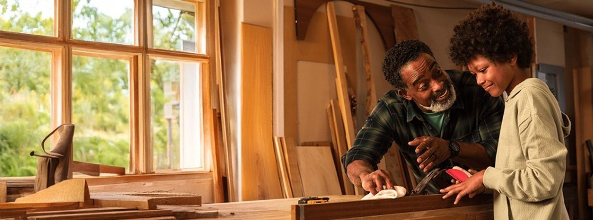 An older man and a young boy smiling as they work together on a woodworking project in a sunlit workshop.
