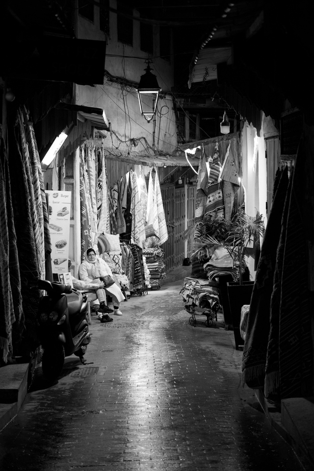 A woman sits outside a carpet shop lit by a street lamp at night