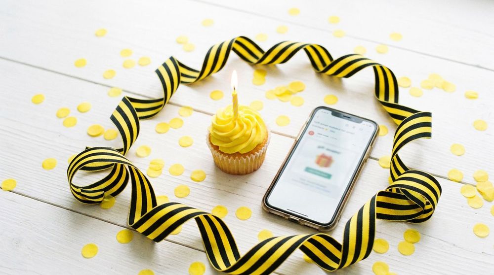 Birthday cupcake with a lit candle beside a phone showing a Vitamin Shots gift card email, with yellow confetti and black and yellow ribbon