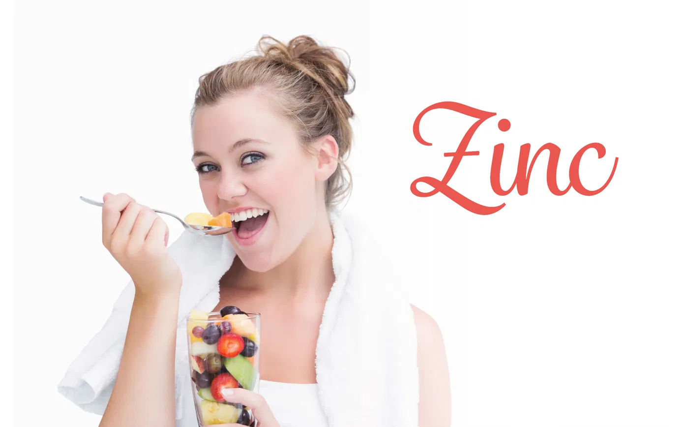 Smiling young woman in workout clothes eating a colourful fruit salad with the word Zinc displayed beside her — illustrating zinc-rich foods and a balanced vegan diet for immune health