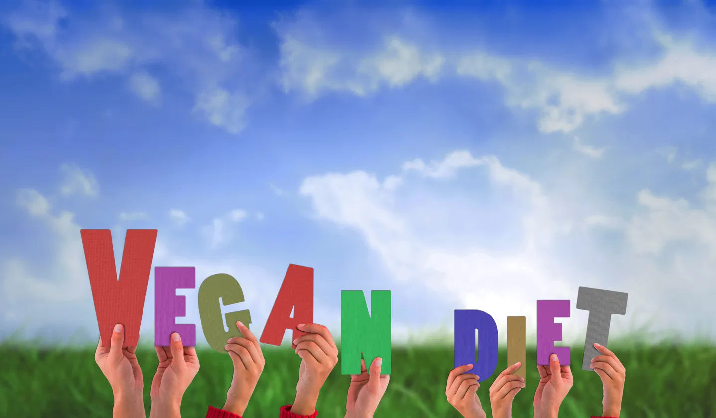 Hands holding up colourful letter cut-outs spelling VEGAN DIET against a blue sky and green grass background — illustrating the vegan lifestyle and plant-based eating community