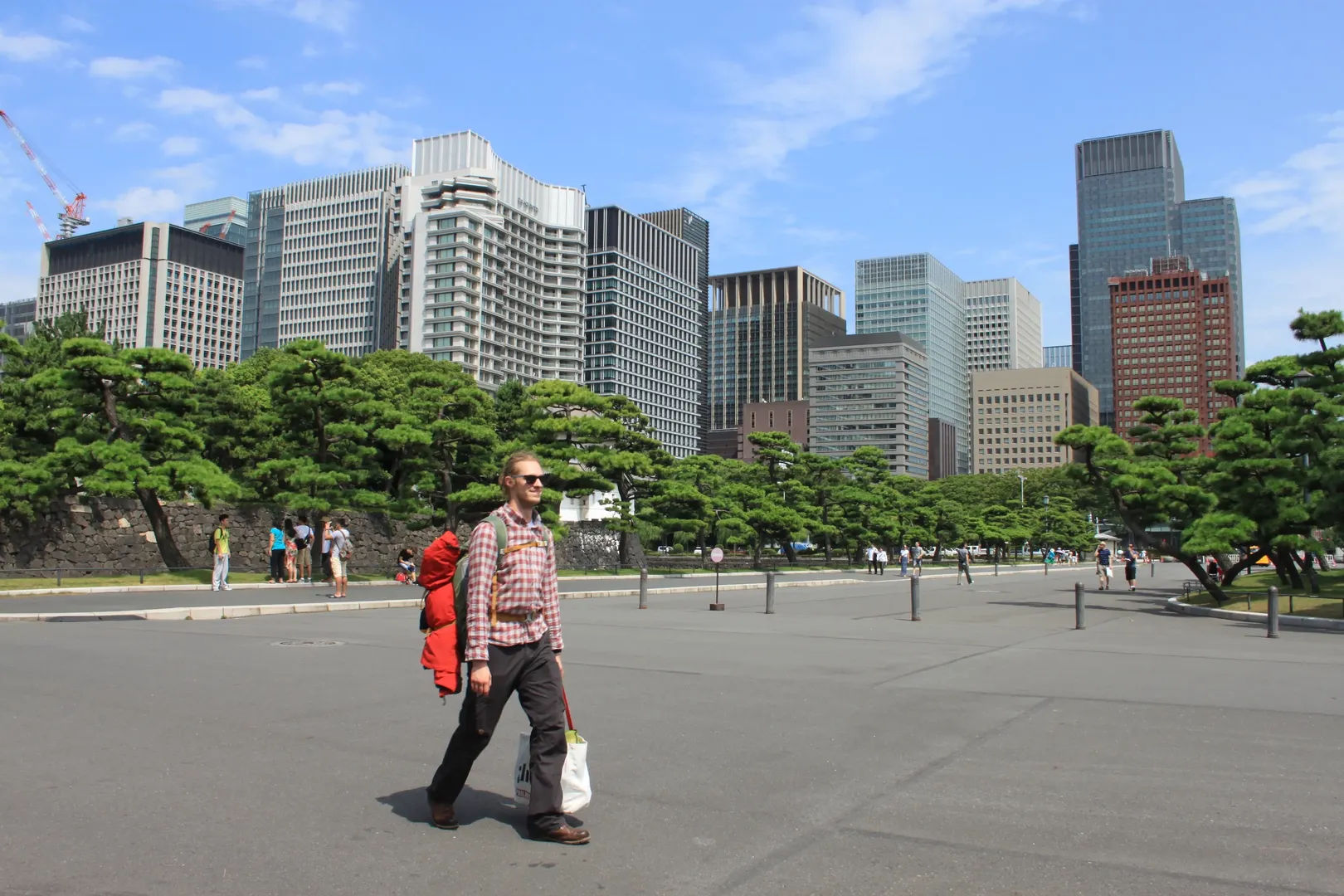 Osoba idąca z plecakiem przez plac przy Pałacu Cesarza z drzewami i wieżowcami Marunouchi w tle, Tokio, Japonia