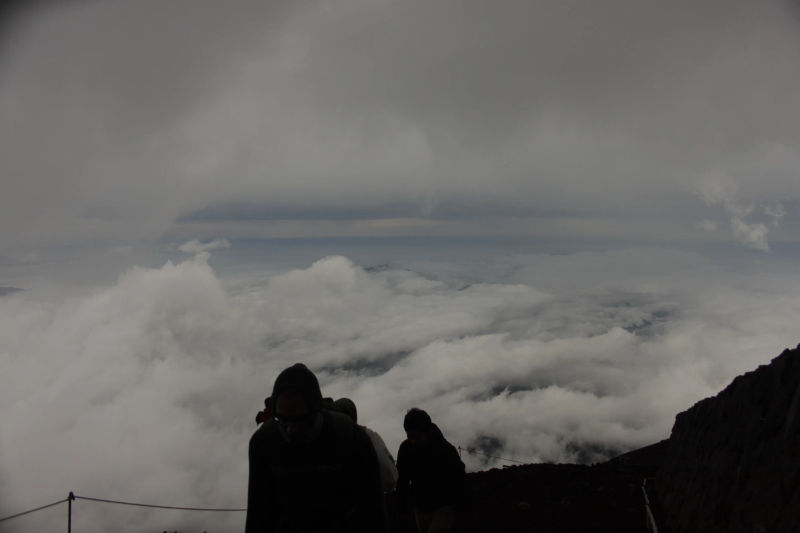 Widok na chmury z wysokości na szlaku górskim na Fuji, z sylwetkami turystów w dolnej części obrazu.
