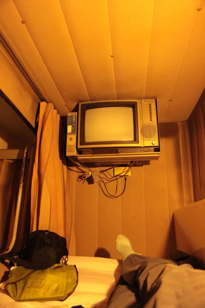 Wnętrze kapsuły w hotelu kapsułowym w Japonii, z widokiem na leżącą osobę i zamontowany na suficie stary telewizor.