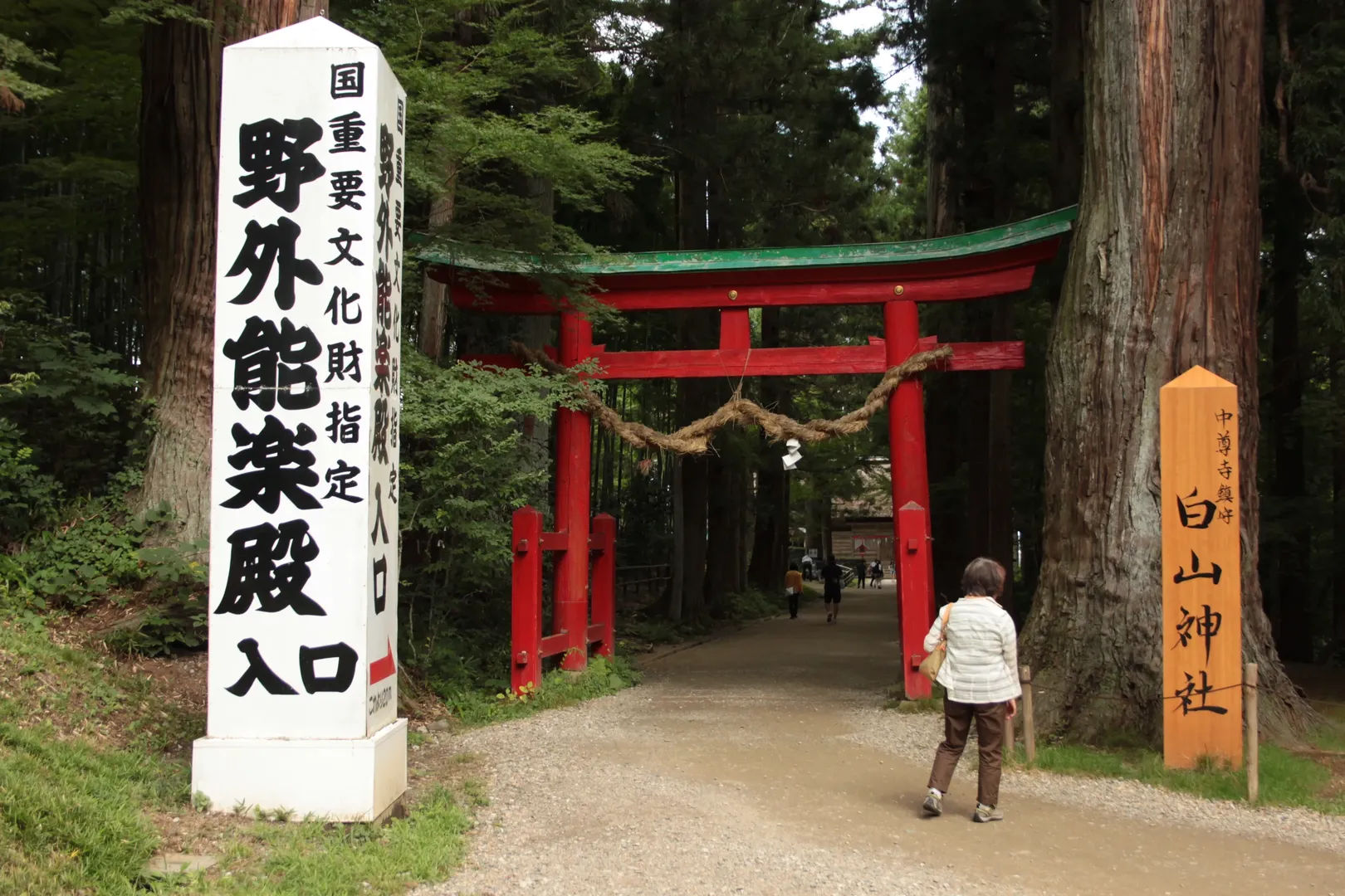 Wejście do świątyni z czerwonym torii i dużym kamiennym znakiem, otoczone drzewami, w kompleksie Chuson-ji w Hiraizumi