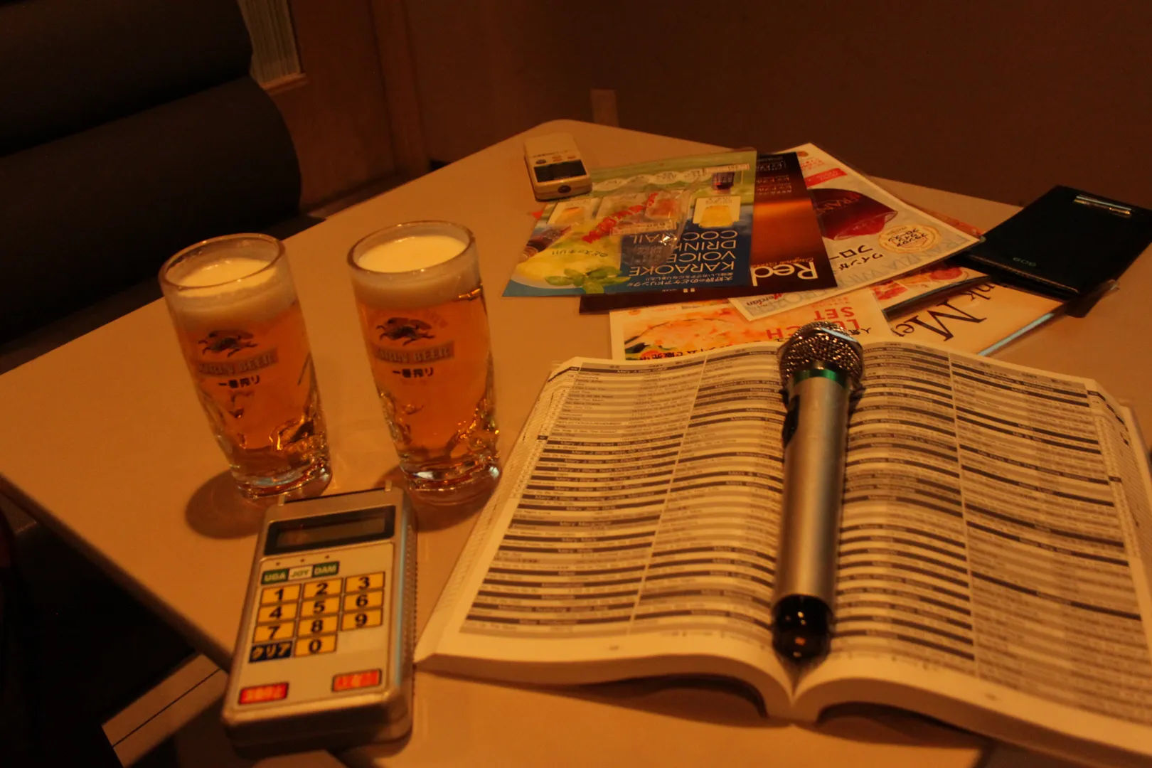 Stolik w pokoju karaoke w Tokio, na którym znajdują się dwa kufle piwa, pilot do obsługi karaoke, mikrofon, otwarta książka z tekstami piosenek oraz różne katalogi i ulotki.