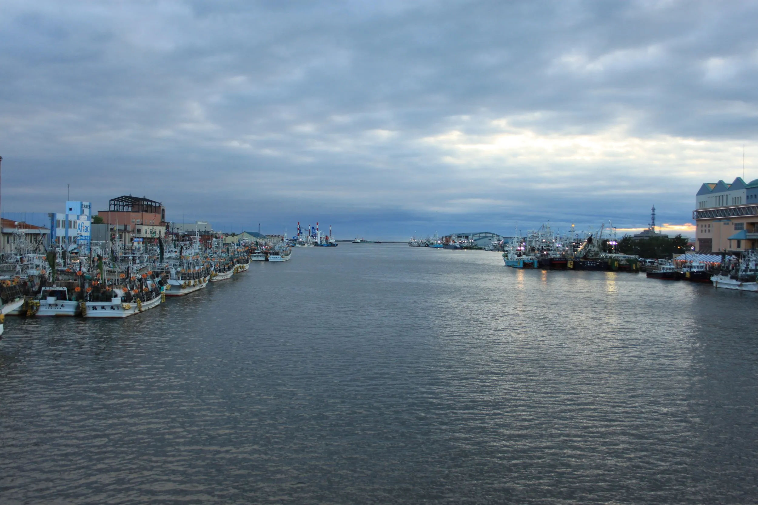 Szeroki widok na port w Kushiro z licznymi zacumowanymi łodziami po obu stronach rzeki, pod pochmurnym niebem