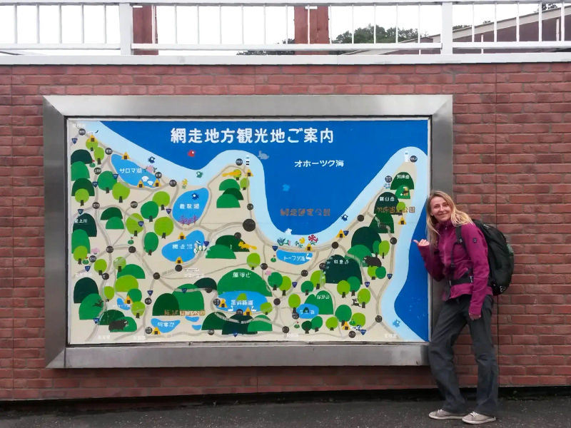 Kobieta z plecakiem stojąca przy mapie z licznymi jeziorami, mapa jest napisana po japońsku'