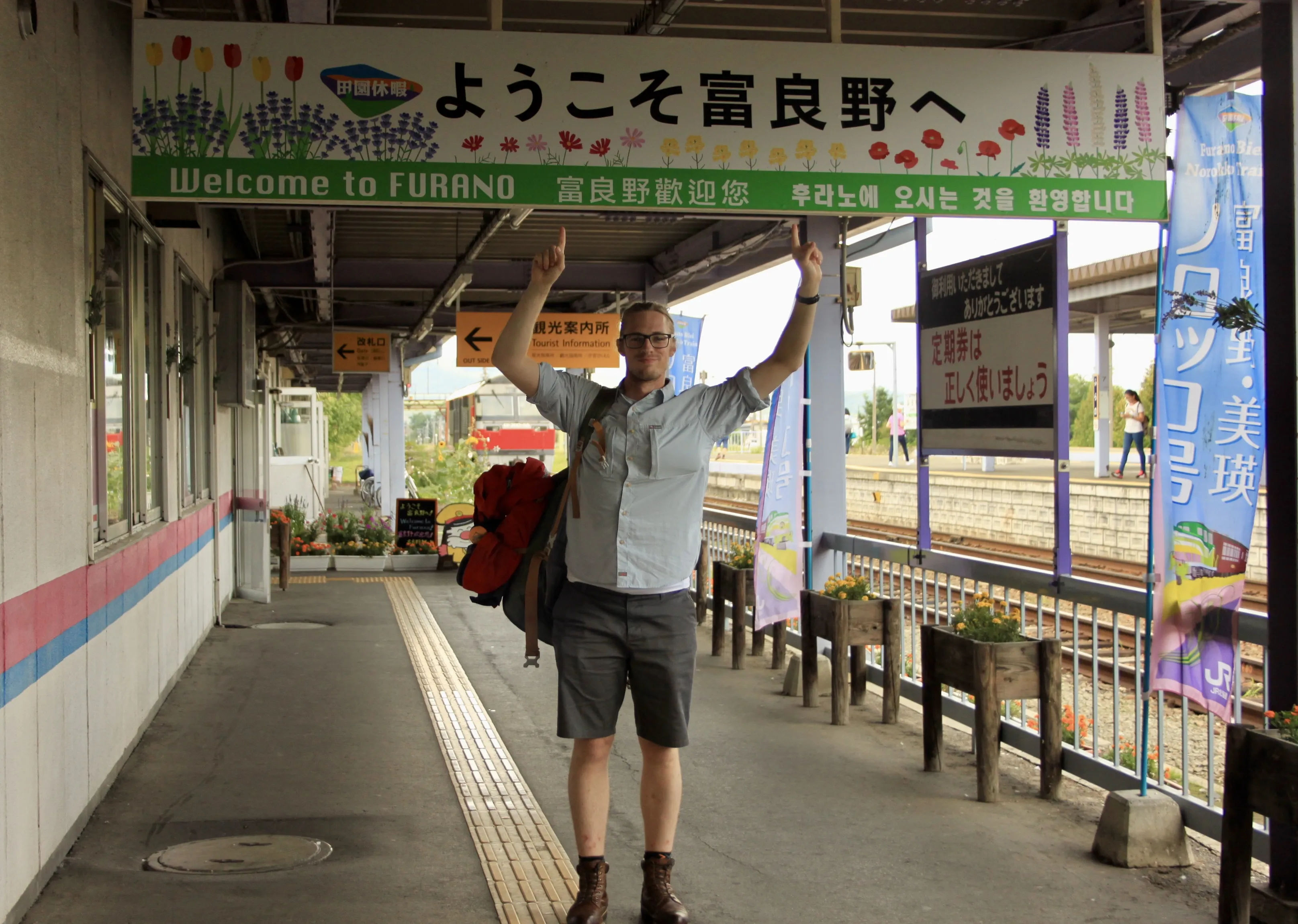 Mezczyzna stojący na stacji kolejowej w Nakafurano, trzymający szyld "Welcome to Furano" wiszący nad nim