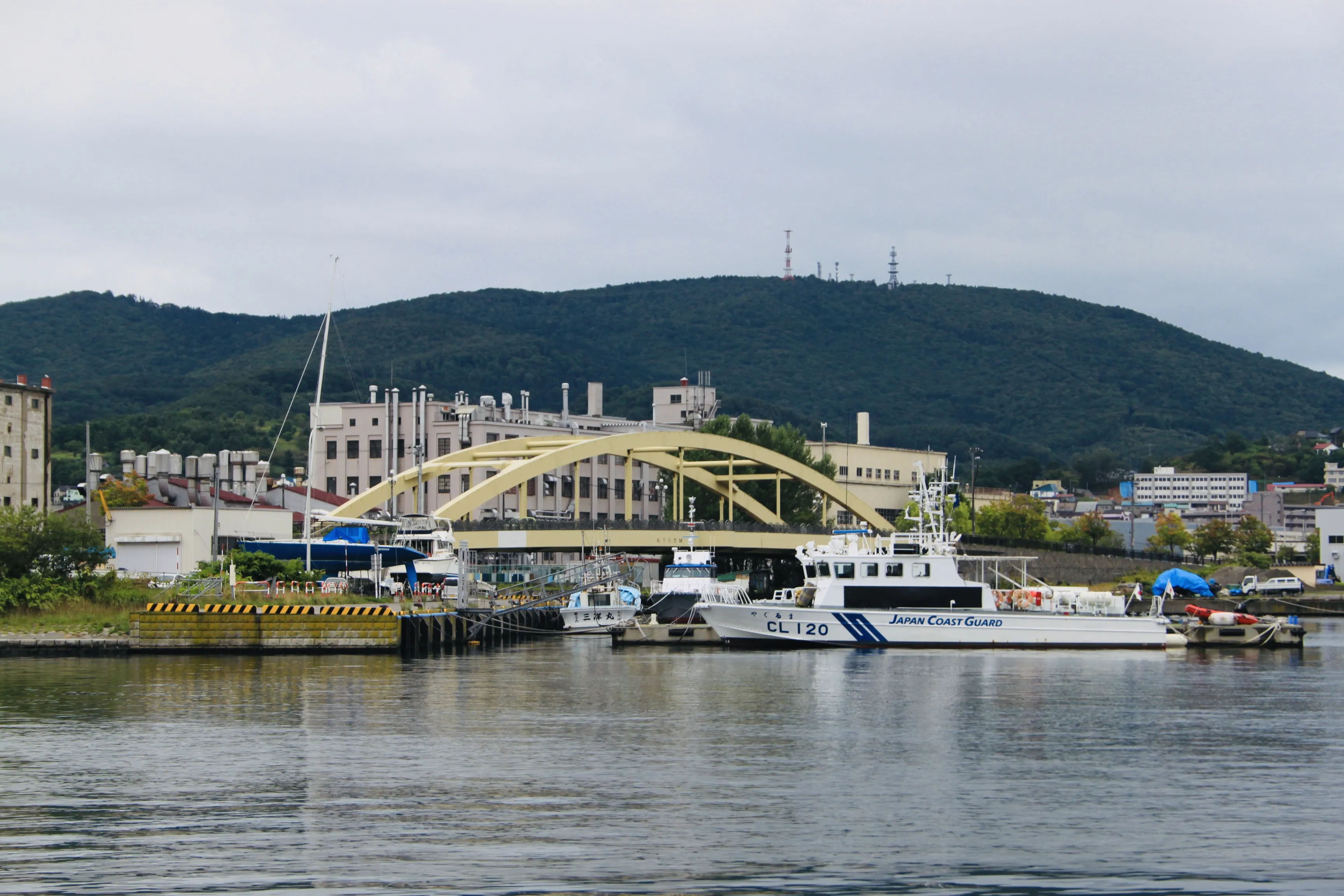 alt="Widok na most w Otaru z łodziami na pierwszym planie i zielonymi wzgórzami w tle