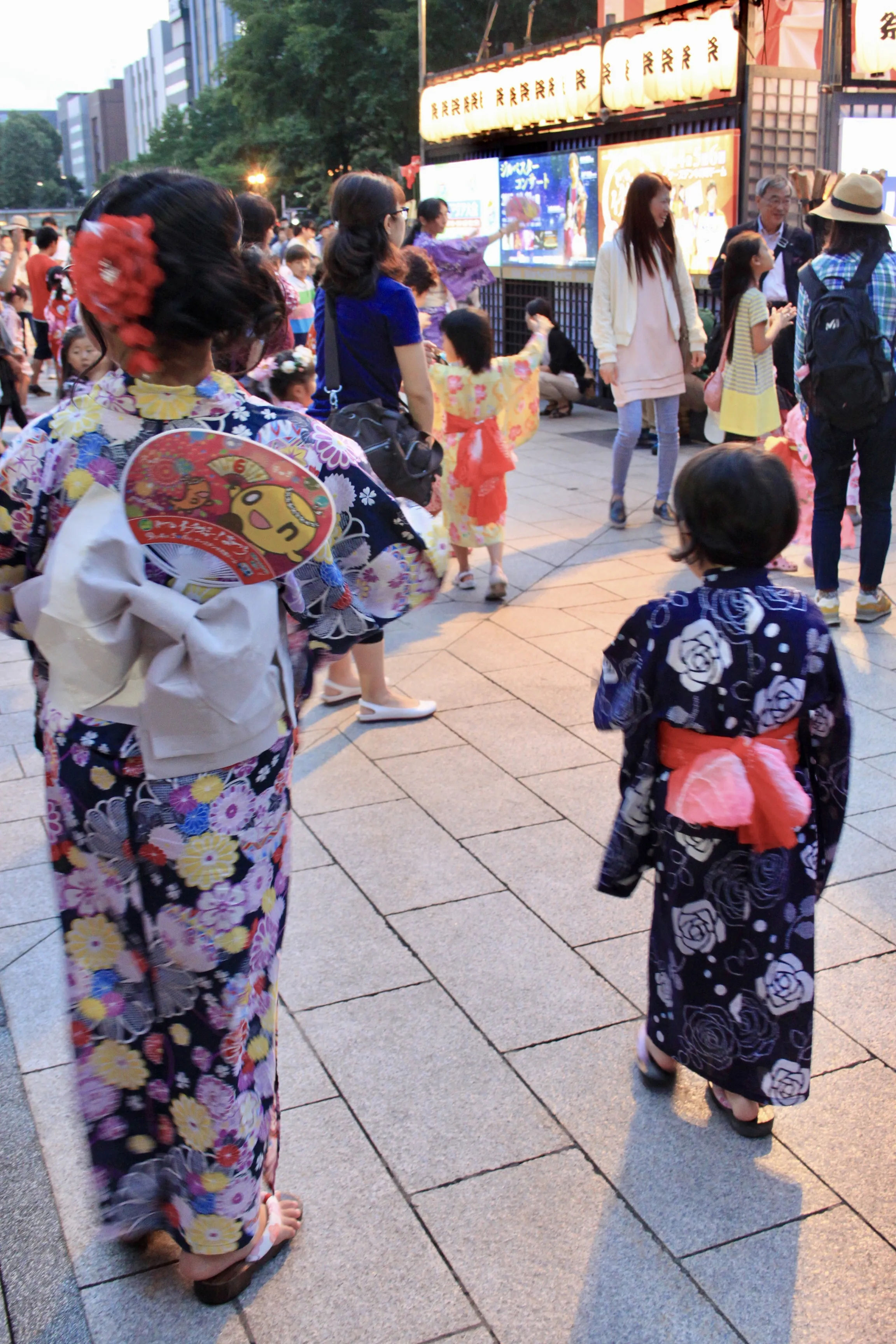 Japonka w kimono i dziewczynka w kimono na pierwszym planie, w tle przechodnie i stoisko z jedzeniem