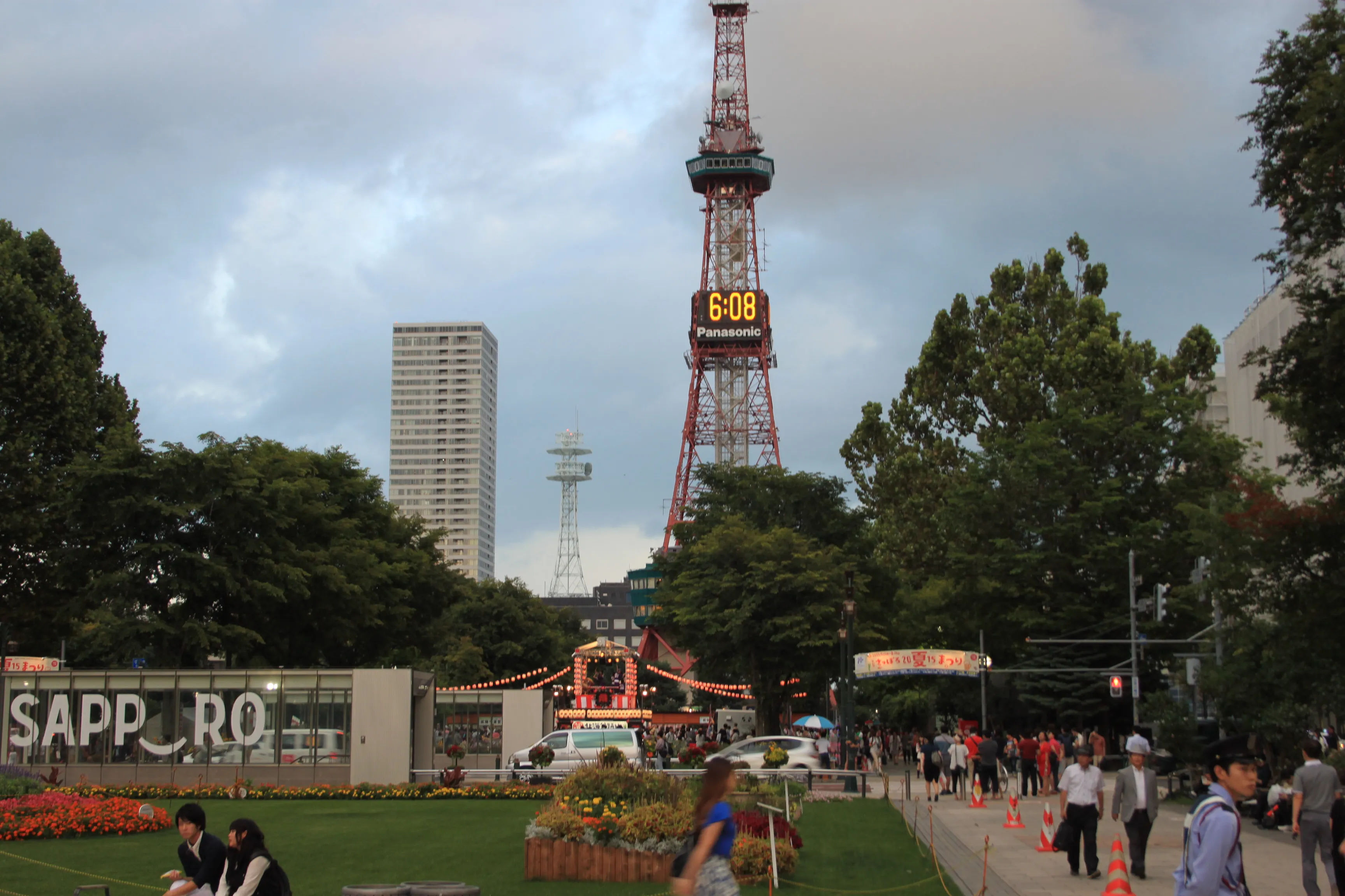 Park Odori w Sapporo z ludźmi zmierzającymi pod scenę Bon Odori, duża wieża telewizyjna z zegarem, zbudowana scena, budynki z napisem Sapporo, wieżowiec w oddali i liczne drzewa