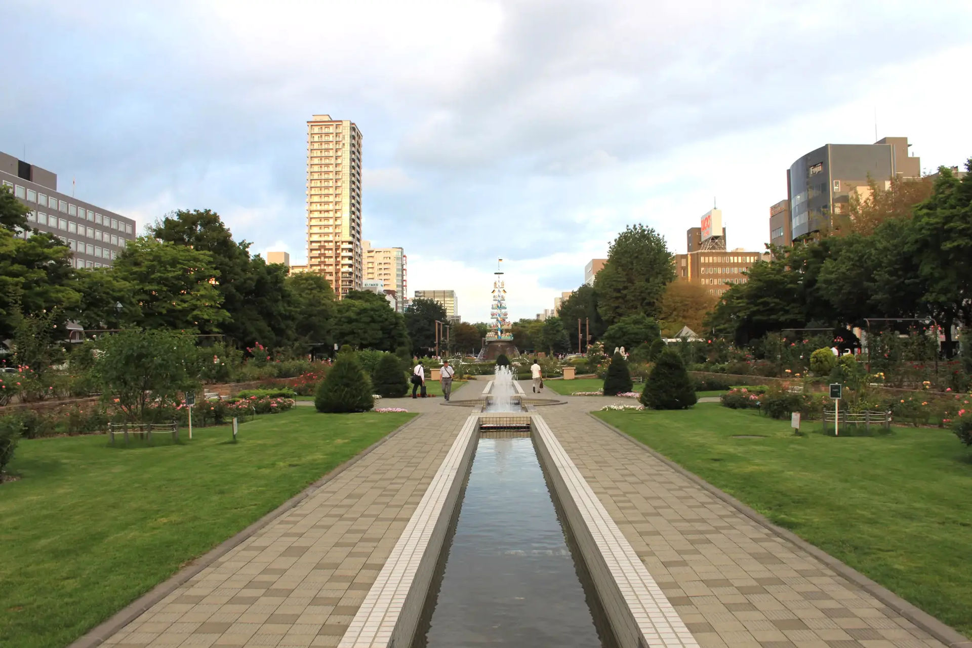 Odori Park w Sapporo z kanałem wodnym, kończącym się konstrukcją przypominającą niemiecki Maibaum, otoczony trawą i budynkami w tle