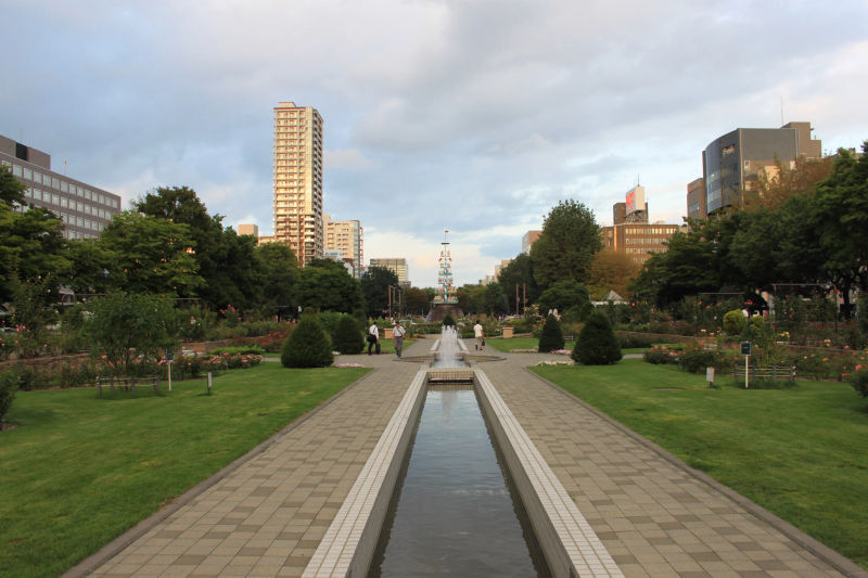 Odori Park w Sapporo z kanałem wodnym, kończącym się konstrukcją przypominającą niemiecki Maibaum, otoczony trawą i budynkami w tle