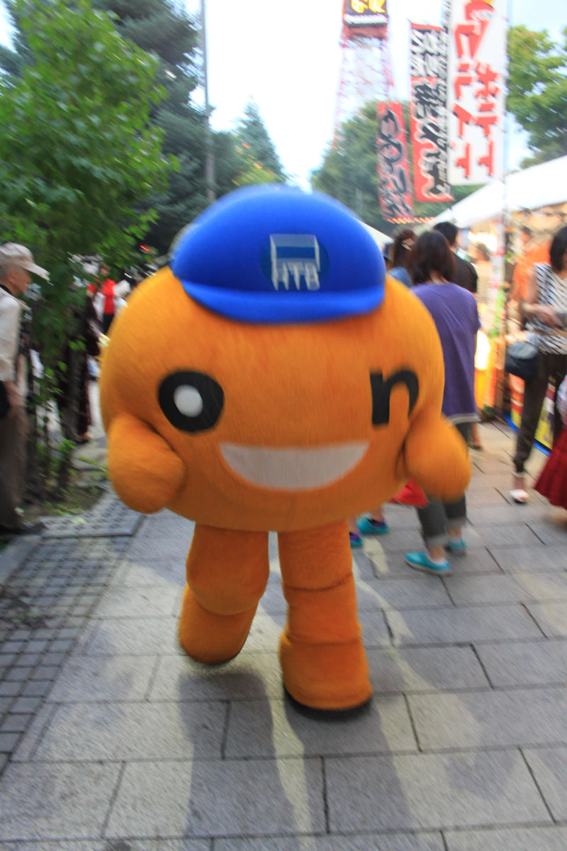 Pomarańczowa maskotka z niebieską czapeczką z napisem HTB na ulicy Parku Odori w Sapporo, z przychodnią w tle