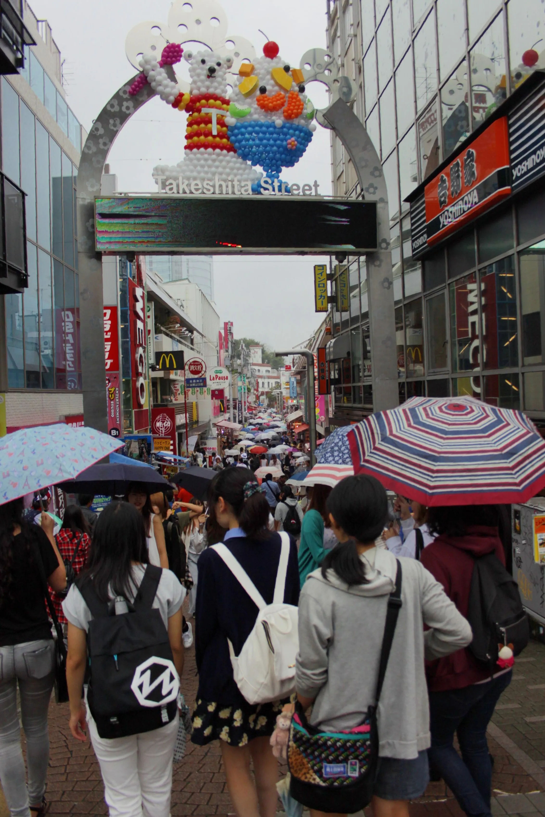Tłum ludzi pod parasolami na ulicy Takeshita Street w Harajuku, z widoczną bramą wejściową udekorowaną balonami.