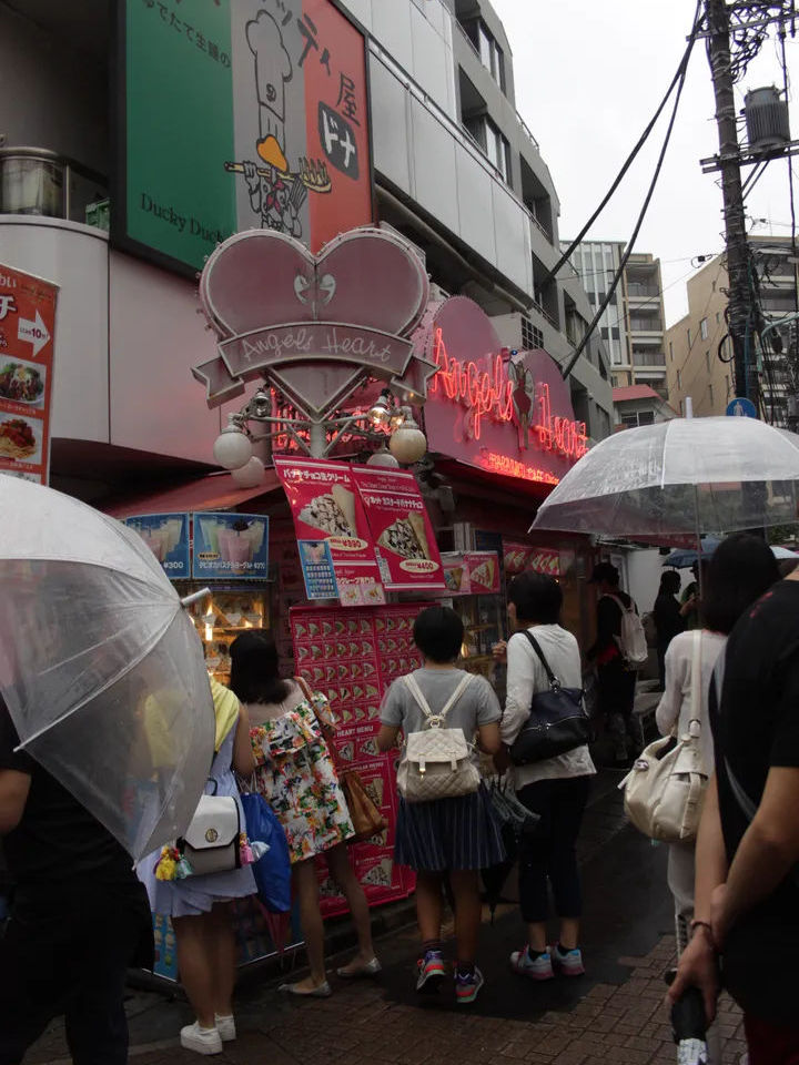 Ludzie stojący przy stoisku na ulicy Takeshita Street w Harajuku, trzymający parasole podczas deszczu.