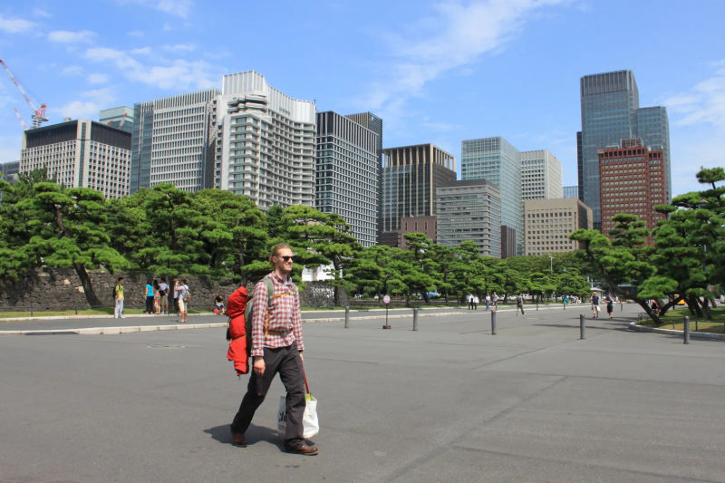 Osoba idąca z plecakiem przez plac przy Pałacu Cesarza z drzewami i wieżowcami Marunouchi w tle, Tokio, Japonia