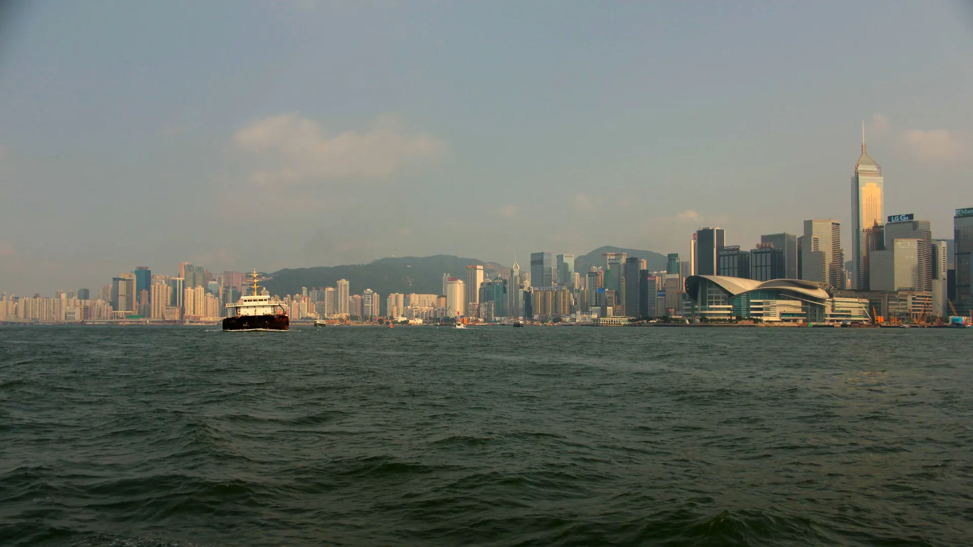 Statek na tle panoramy Hongkongu, z dużymi wieżowcami i górami w oddali