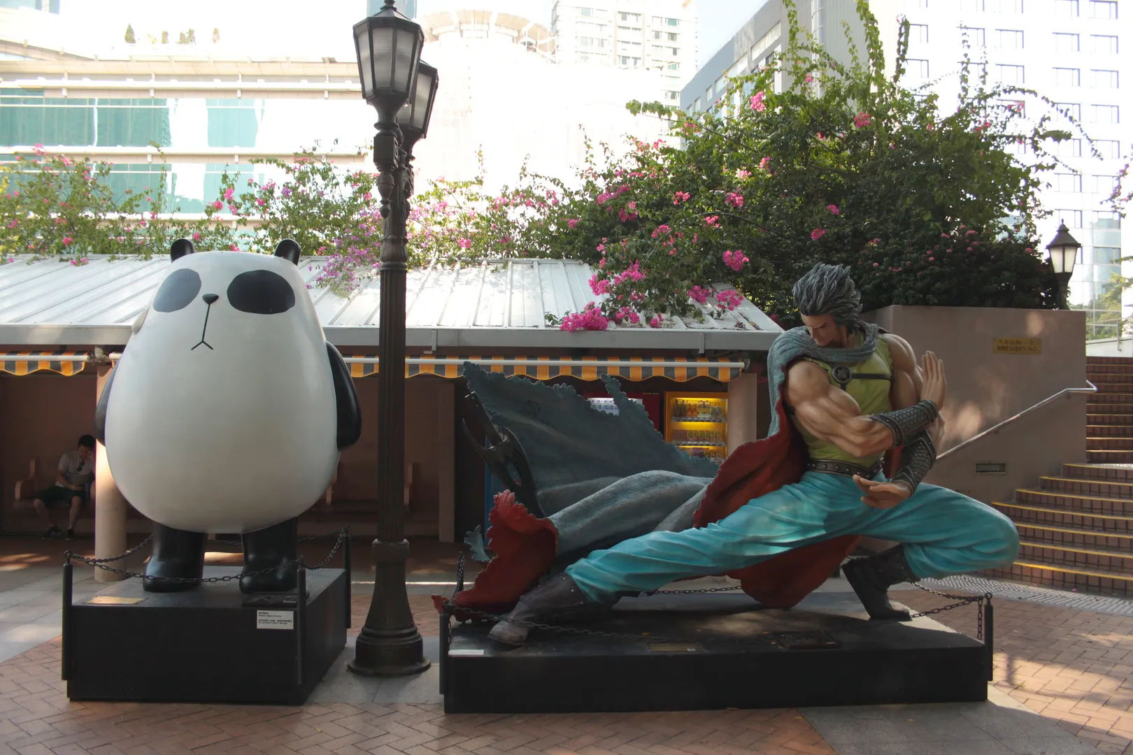 Postacie z kreskówek stojące wzdłuż deptaku w Tsim Sha Tsui, Hongkong, otoczone palmami i wysokimi budynkami.