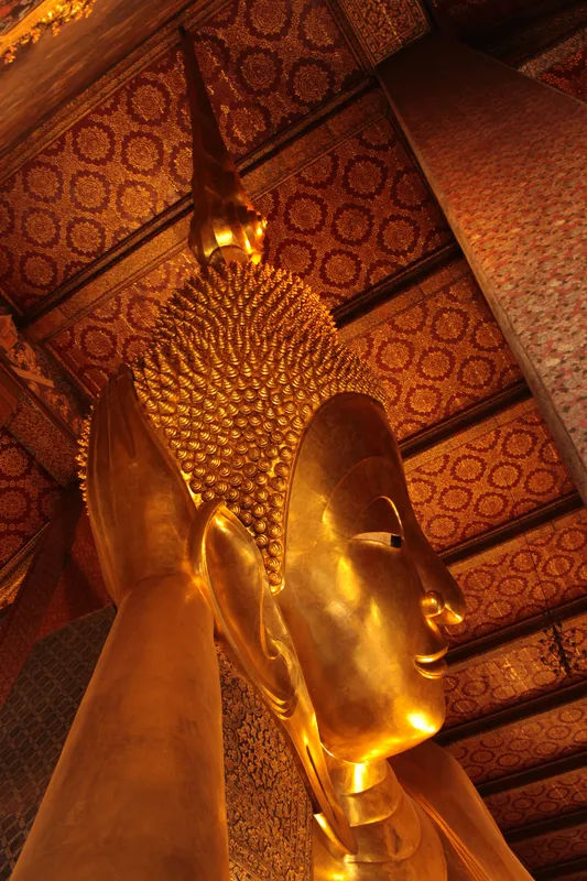 Złocisty posąg Buddy w Wat Pho w Bangkoku, z misternymi detalami na tle złotej ściany.