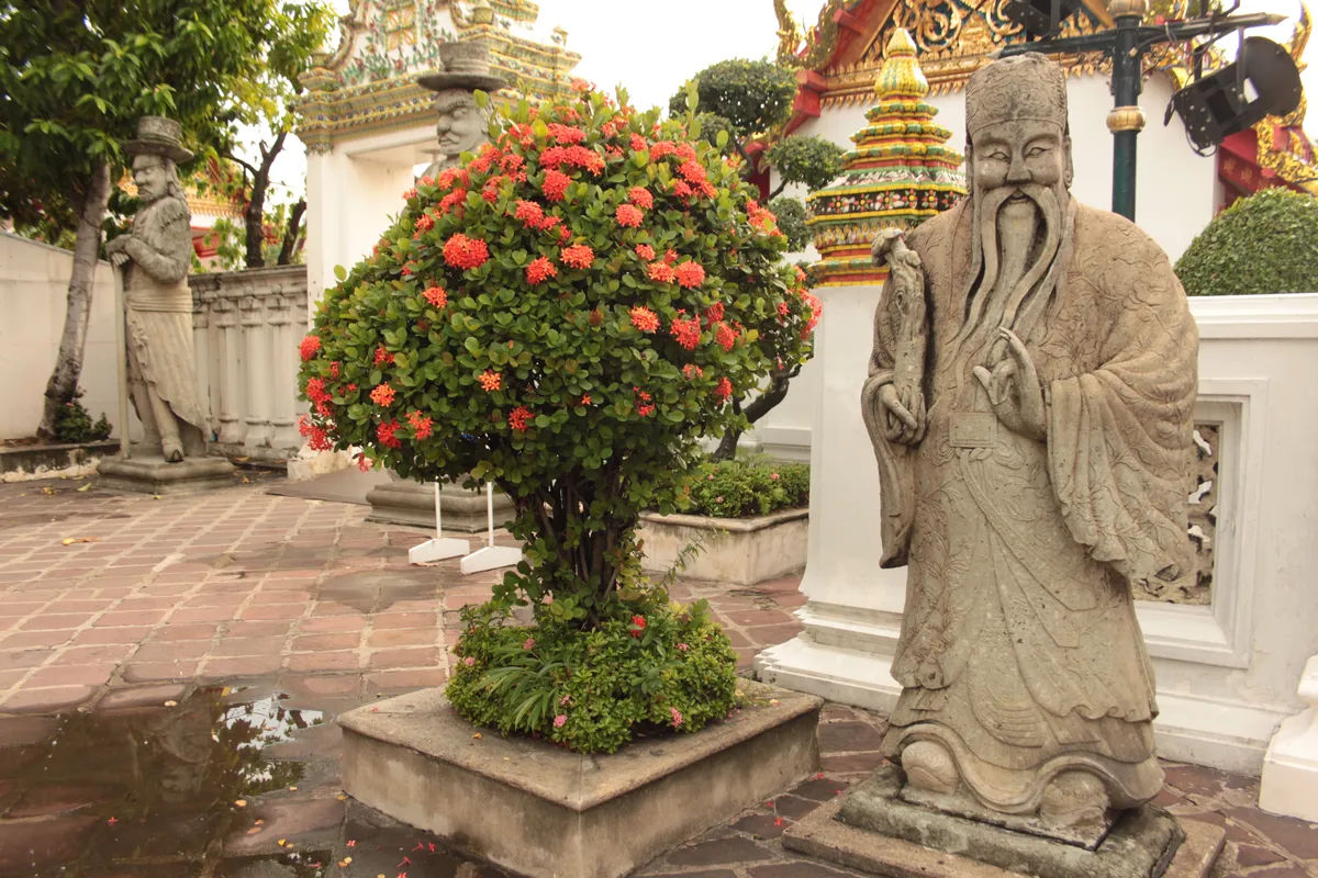 Posąg mędrca w Wat Pho w Bangkoku, otoczony roślinnością i tajską architekturą.