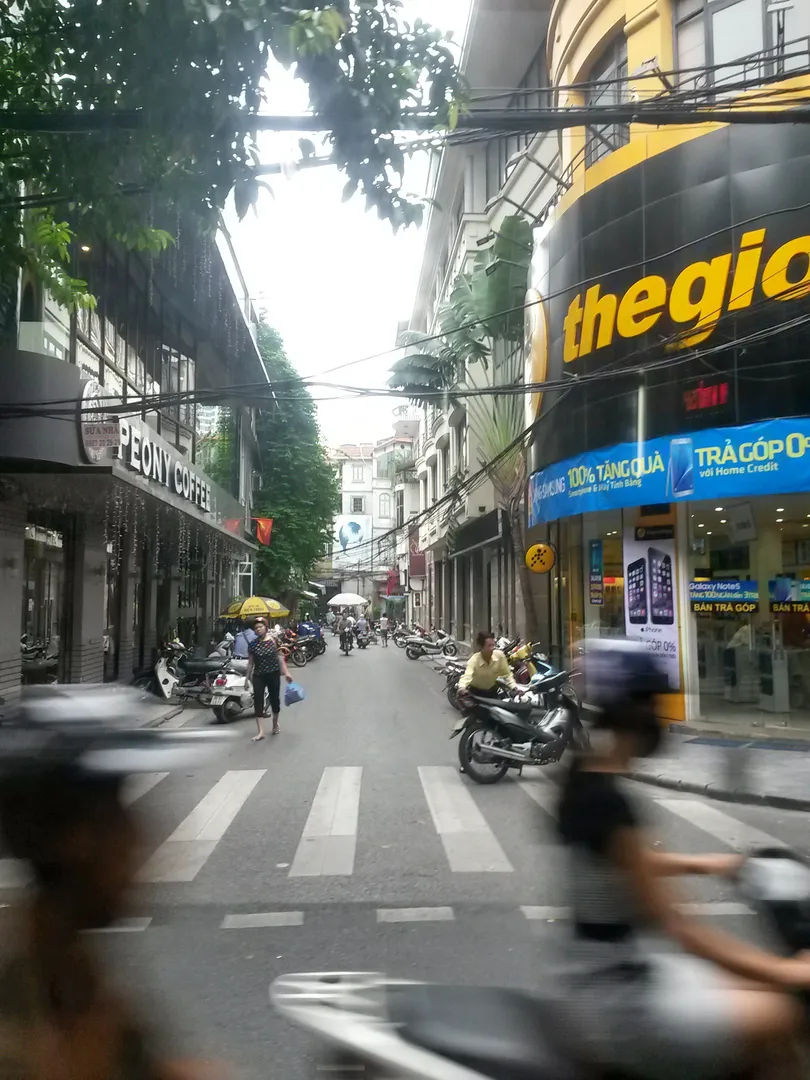 Ruchliwa ulica w Hanoi z motocyklistami i pieszymi, otoczona nowoczesnymi budynkami i sklepami.